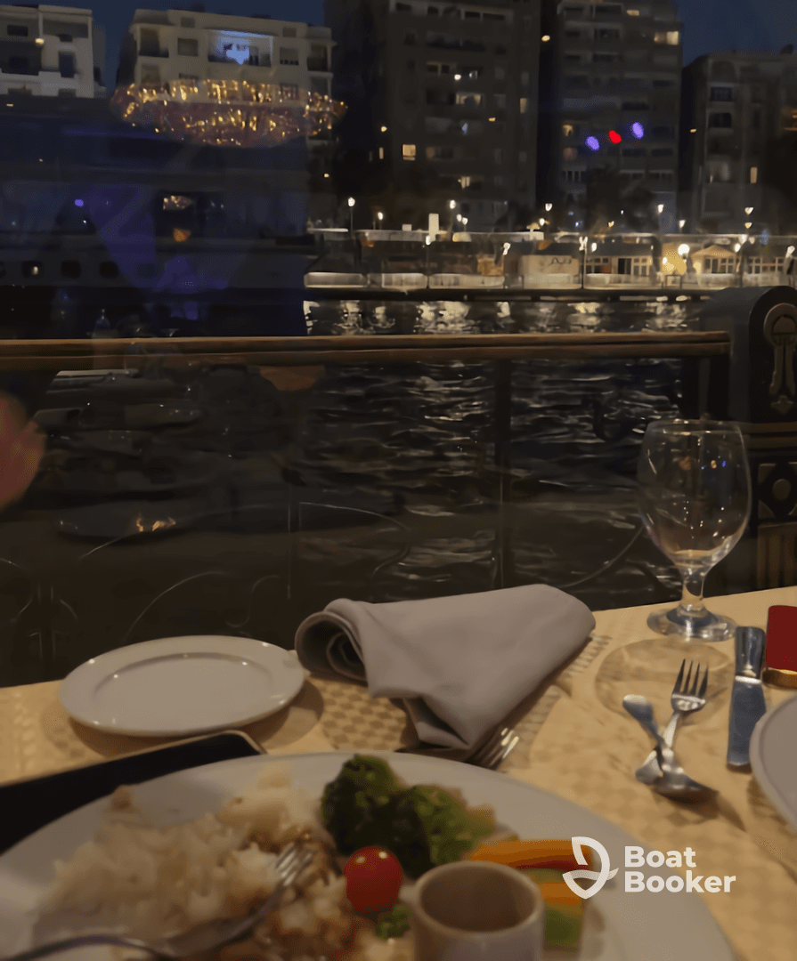 A beautifully set dining table on a boat, featuring a delicious meal with a view of the waterfront at night — perfect for a romantic dinner or special occasion.