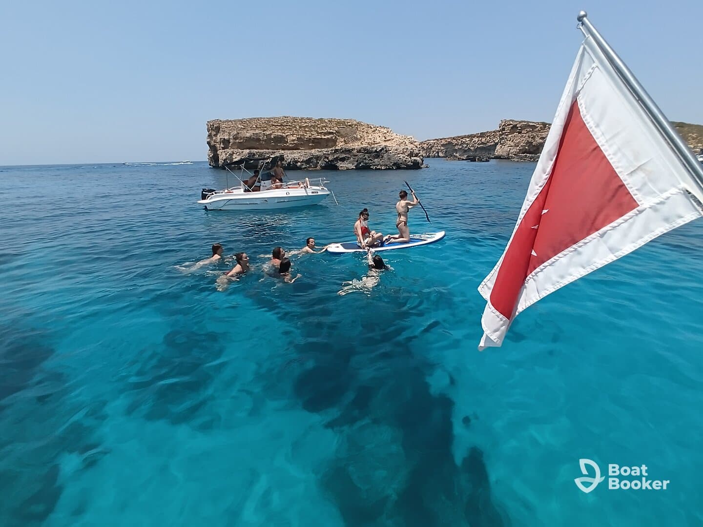 A lively group enjoys paddleboarding and swimming in crystal-clear waters, with a scenic backdrop of rocky cliffs — perfect for summer adventures.