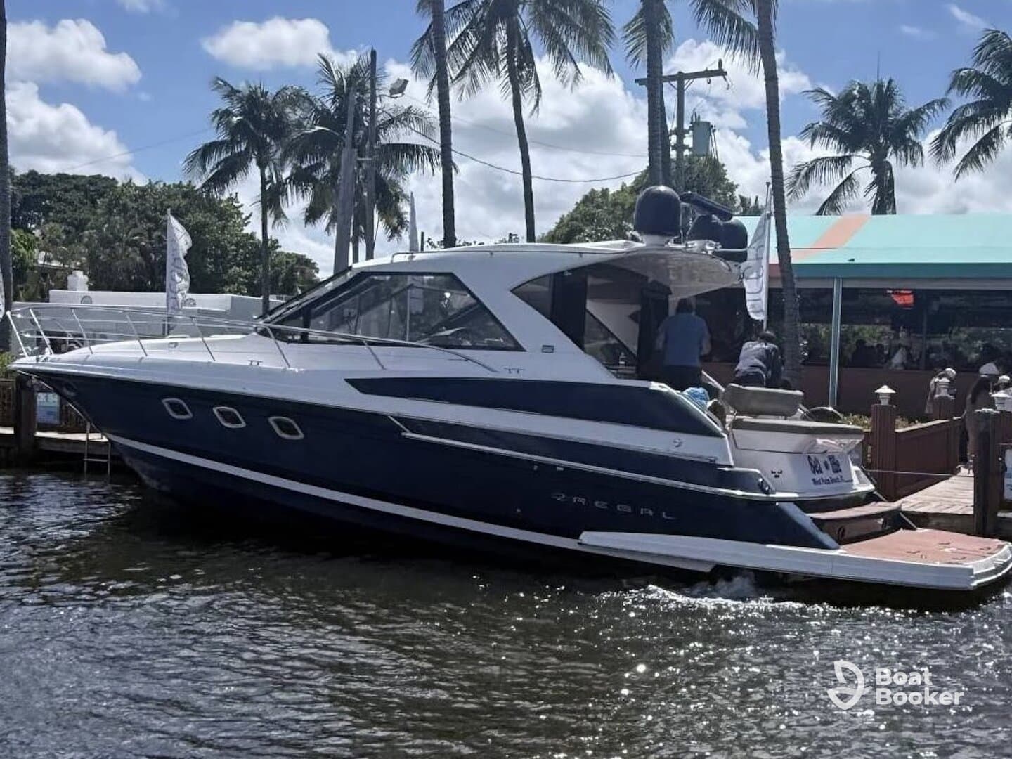 A Regal yacht is docked, showcasing its sleek design against a backdrop of palm trees and a lively waterfront scene.