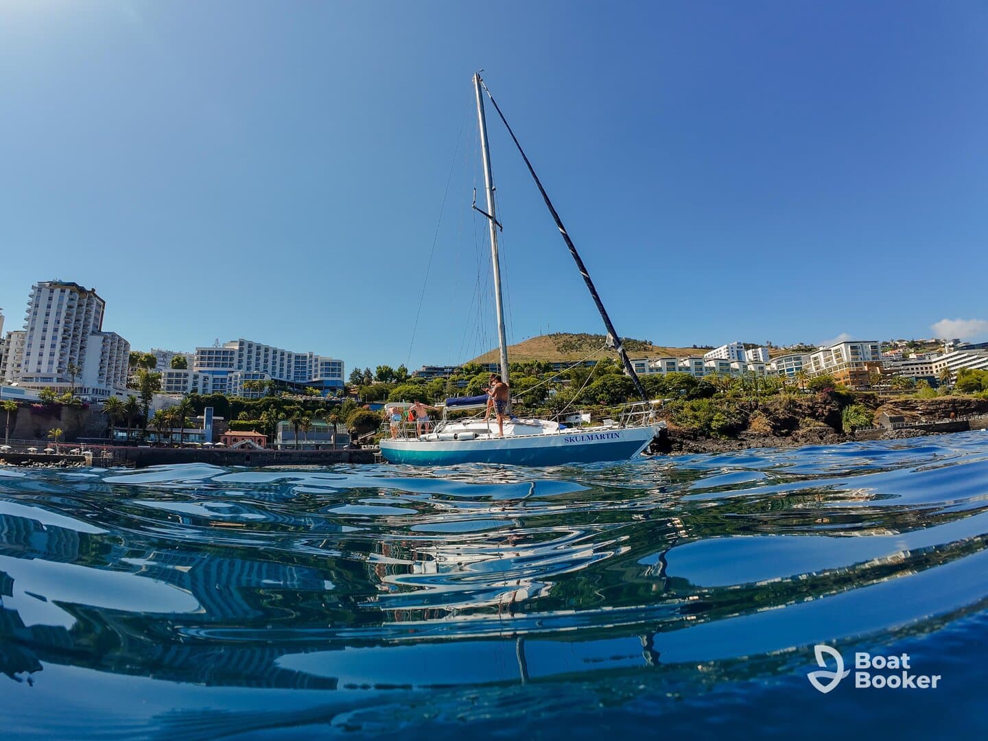 A picturesque sailboat named 'SKOLMARTIN' is anchored near a vibrant coastal area, showcasing the beauty of the surrounding landscape and clear waters.