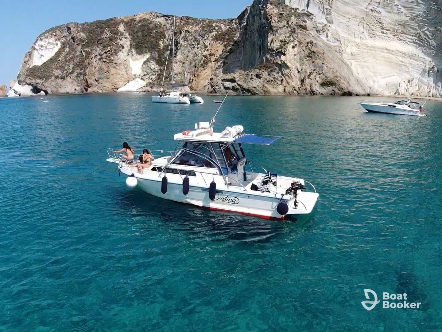 Two women relax on the bow of a boat in crystal-clear waters, surrounded by stunning coastal scenery — a perfect moment for a summer getaway.