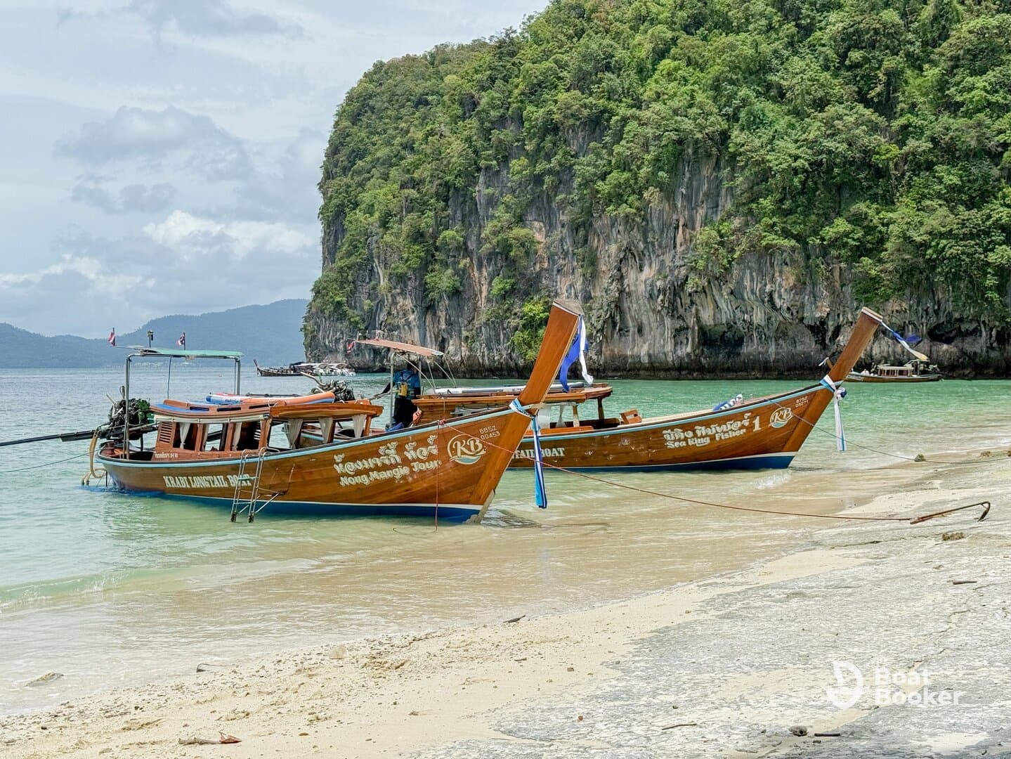 Two traditional longtail boats are docked on a sandy beach, surrounded by lush greenery and rocky cliffs, showcasing a serene coastal scene.