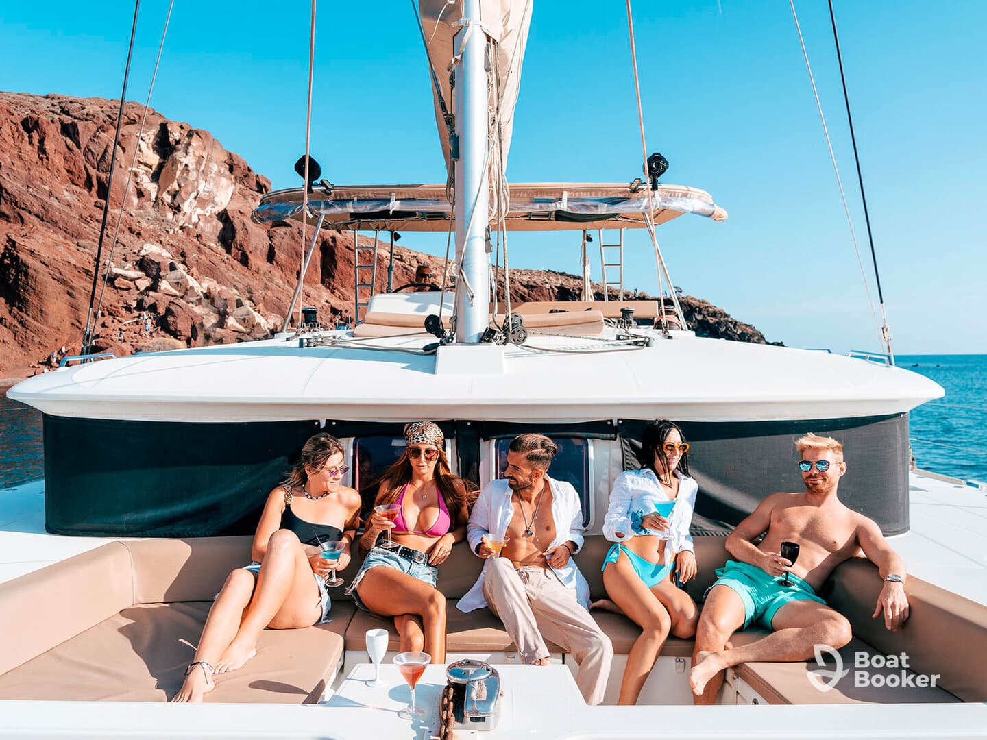 A vibrant group enjoys cocktails and sunbathing on a catamaran, surrounded by stunning coastal views — perfect for a fun day on the water.
