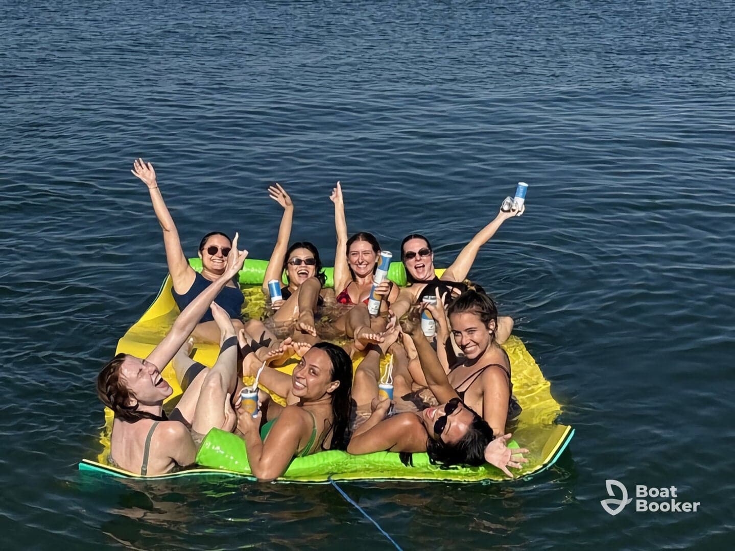 A lively group of friends enjoys a fun day on a floating mat in the water, celebrating with drinks and laughter — perfect for summer parties and memorable outings.