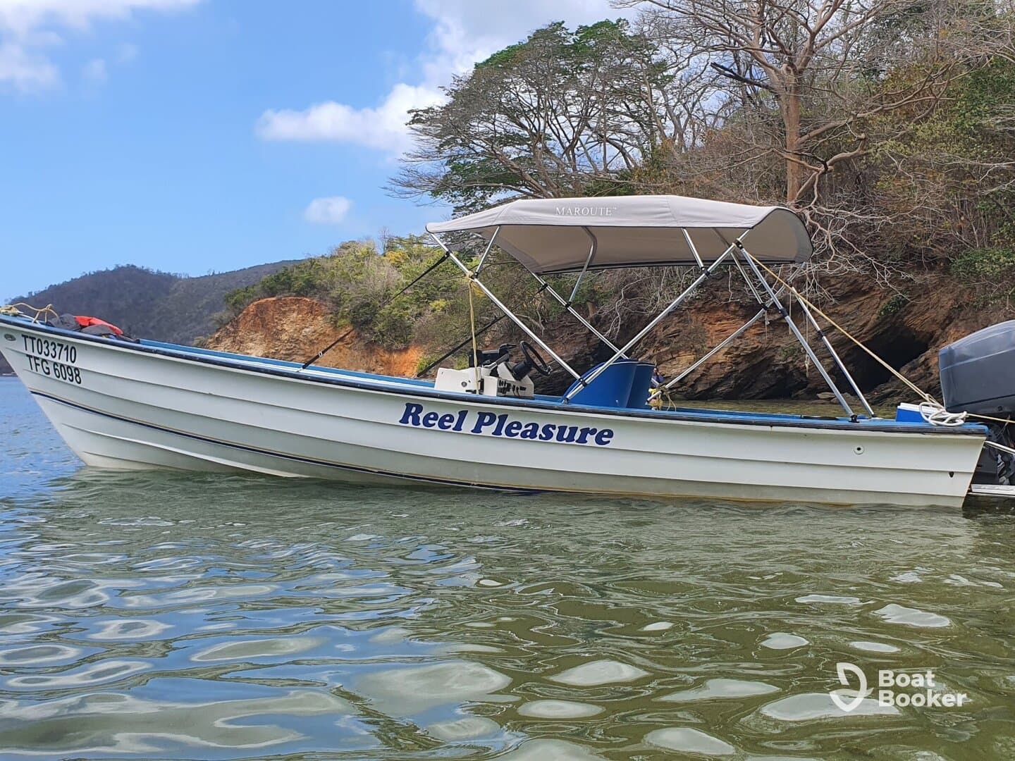 A picturesque view of the 'Reel Pleasure' boat anchored in calm waters, surrounded by lush greenery and rocky cliffs, perfect for a day of fishing or leisure on the water.