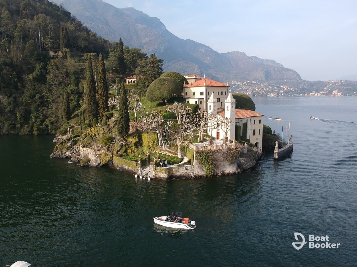 A picturesque view of a stunning lakeside villa surrounded by lush greenery and mountains, with a small boat navigating the serene waters — a perfect representation of scenic beauty and tranquility.