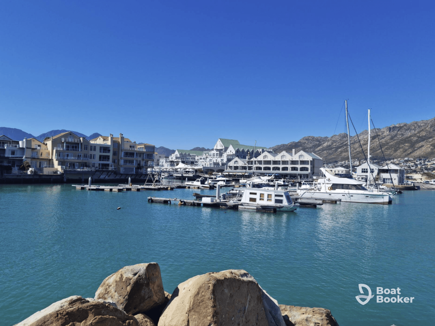 A picturesque view of a marina featuring various yachts and catamarans docked against a backdrop of scenic mountains and clear blue skies.