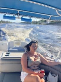 A woman enjoys a sunny day on a boat, smiling and holding a drink while cruising through the water — a perfect moment for relaxation and fun on the water.