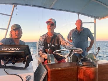 Three individuals are actively engaged at the helm of a boat during sunset, showcasing the excitement of navigating the open waters.