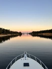 A serene view from the bow of a boat, capturing a tranquil waterway at sunset, framed by lush trees and a distant bridge.