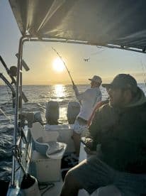 Two fishermen enjoy a sunset fishing trip, with one reeling in a catch while the other relaxes on the boat — a perfect blend of adventure and tranquility on the water.
