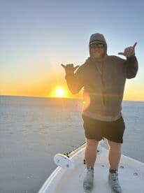 A man enjoys a sunset on a boat, striking a fun pose as the sun sets over the horizon — a perfect moment for beach and boating enthusiasts.