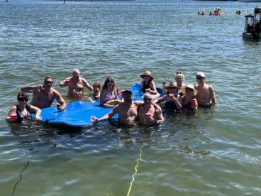 A lively group enjoys a fun day in the water, relaxing on a floating mat with drinks in hand — perfect for summer parties and celebrations.