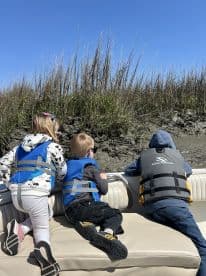Three children in life jackets enjoy a day on the boat, peering over the edge to explore the natural surroundings — a perfect moment of adventure on the water.
