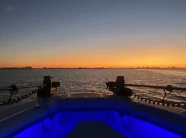 A serene sunset view from the bow of a boat, showcasing the tranquil waters and city skyline in the distance — perfect for evening cruises.