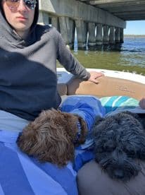 A young man relaxes on a boat with two dogs, enjoying a serene day on the water under a bridge. Perfect for pet-friendly boating adventures.