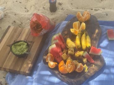 A vibrant spread of fresh fruit, guacamole, and chips laid out on a sandy beach, perfect for a relaxing day on the water or a picnic by the shore.