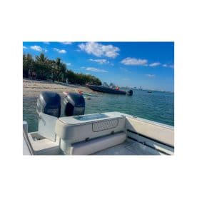 A view of a speedboat with twin 300 HP engines, anchored near a sandy beach with people enjoying the sun and water activities in the background.