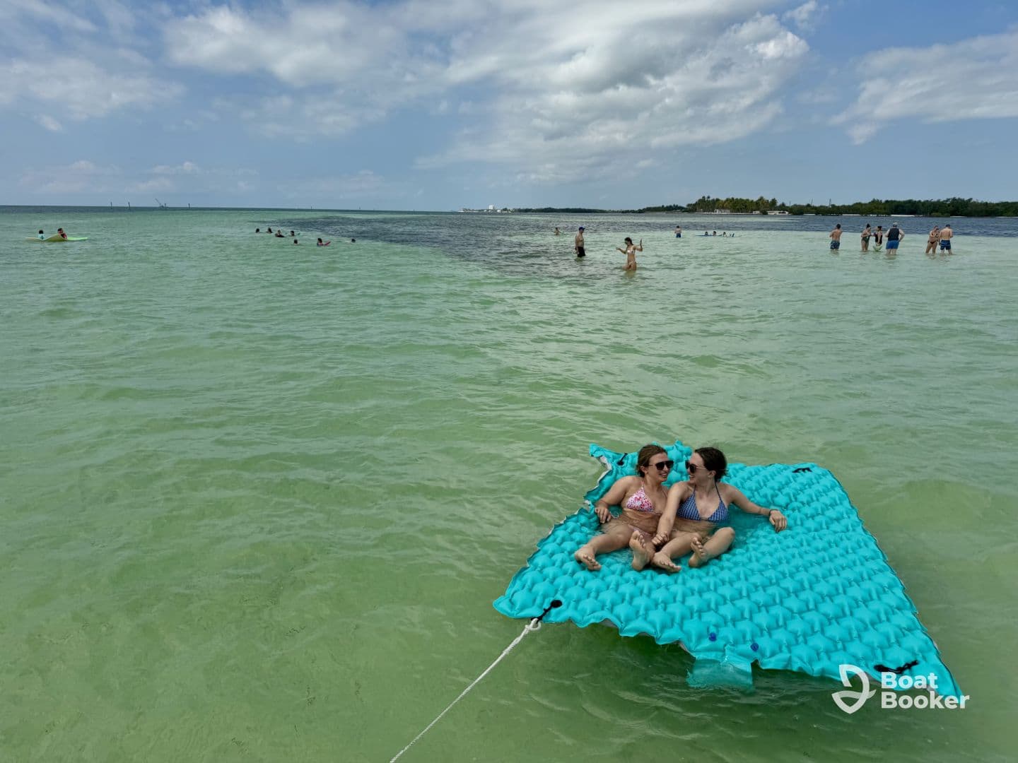 Two friends relax on a floating mat in shallow waters, surrounded by others enjoying a sunny beach day — a perfect scene for summer fun and relaxation.