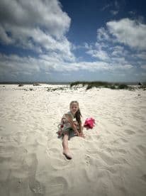 A young girl enjoys a sunny day on the beach, playing in the sand and smiling for the camera — a perfect moment of childhood joy by the water.