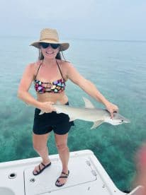 A woman proudly holds a shark while standing on the edge of a boat in clear waters, showcasing an exciting wildlife encounter.