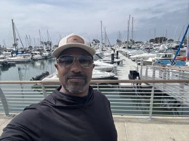 A man takes a selfie at a bustling marina filled with various boats, capturing the vibrant atmosphere of a day on the water.