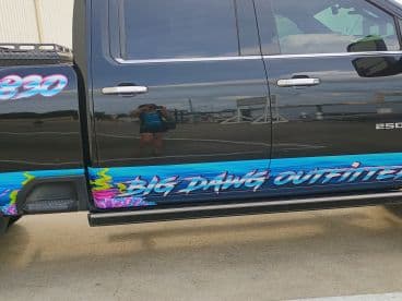 A close-up shot of a truck featuring vibrant graphics and the name 'Big Dawg Outfitters.' The reflection of a person is visible, adding a personal touch to this vehicle's branding.