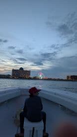 A child sits on the bow of a boat, gazing at a vibrant cityscape with a colorful ferris wheel at sunset — a perfect moment for family outings on the water.