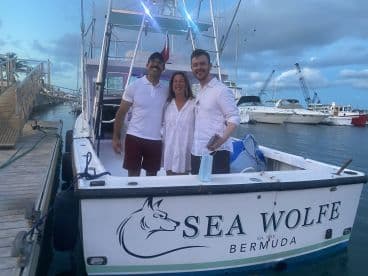 A cheerful group poses on the deck of the 'Sea Wolfe' boat in Bermuda, capturing a memorable moment at the marina.