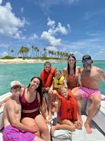 A joyful family poses on a boat, enjoying a sunny day on the water with a beautiful beach backdrop — perfect for memorable summer adventures.