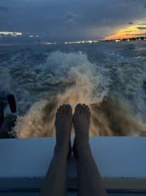 A serene evening shot featuring feet resting on the edge of a boat, capturing the beautiful sunset and the wake of the water — perfect for those enjoying a relaxing boat ride.