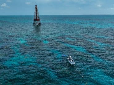 An aerial view captures a boat anchored near a lighthouse, with snorkelers exploring the vibrant waters — a perfect scene for adventure seekers in a tropical paradise.
