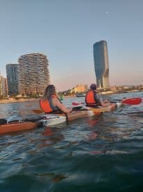 Two kayakers paddle along the waterfront, enjoying the sunset with a stunning city skyline in the background — a perfect urban adventure on the water.