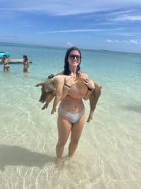 A woman joyfully holds a pig while standing in shallow waters, showcasing a unique wildlife interaction in a tropical setting.