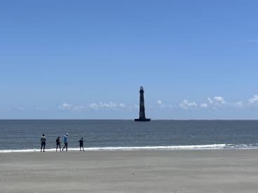 A family strolls along the beach with a picturesque lighthouse in the background, capturing a serene moment by the ocean.