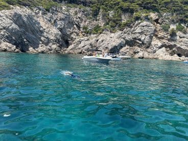 A swimmer enjoys snorkeling in crystal-clear waters near a rocky coastline, with boats anchored nearby, showcasing a perfect day of water activities.