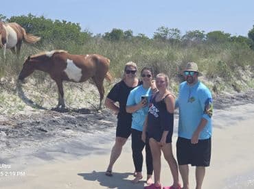 A group of friends poses on the beach with wild horses grazing nearby, capturing a unique wildlife encounter in a picturesque setting.