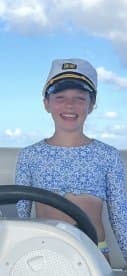 A cheerful child wearing a captain's hat smiles while at the helm of a boat, capturing the joy of a fun day on the water.