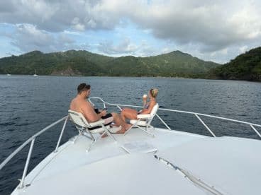 A couple enjoys drinks while relaxing on the bow of a yacht, surrounded by stunning coastal scenery — a perfect moment for unwinding on the water.