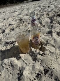 A refreshing cocktail and a bottle of Kalik Radler Lychee sit on the sandy beach, perfect for enjoying a sunny day by the water.