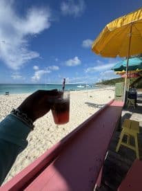 A refreshing cocktail enjoyed on the beach, with a stunning view of the turquoise waters and sailboats in the background — perfect for a relaxing day by the shore.