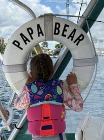 A young girl in a colorful life jacket gazes out from the boat, with a life ring labeled 'PAPA BEAR' in view — capturing a moment of adventure on the water.