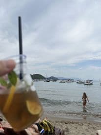 A refreshing cocktail in hand while enjoying a beach day, with a glimpse of boats in the background and a swimmer wading into the water — perfect for a relaxing getaway.
