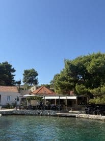 A picturesque waterfront scene featuring a restaurant with outdoor seating, surrounded by lush greenery and clear waters, perfect for enjoying a meal by the sea.