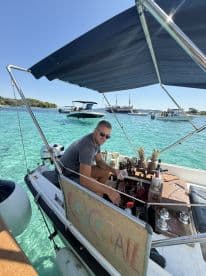 A bartender prepares cocktails on a boat, showcasing a vibrant bar setup amidst clear turquoise waters — perfect for refreshing drinks on a sunny day.