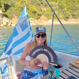 A woman enjoys a sunny day on a boat in Greece, holding a fan with a humorous message while sipping a cocktail — perfect for a fun day on the water.