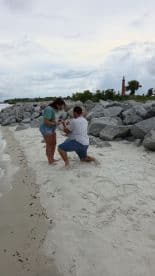 A romantic beach proposal captures a couple's special moment, set against a scenic backdrop with a lighthouse in view.
