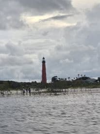 A blurry image capturing a lighthouse with some people in the foreground, set against a cloudy sky. The scene lacks clarity, making it difficult to discern details.