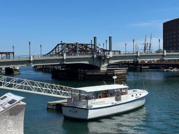 A scenic view of the 'ATLAS' boat docked in Boston, showcasing the vibrant waterfront and nearby bridge.