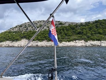 A scenic view of a Croatian flag flying on a boat, with a backdrop of lush green hills and rocky coastline, showcasing the beauty of Croatia's coastal landscape.